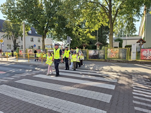Na zdjęciu widoczne dzieci, które przez jednię po przejściu dla pieszych przeprowadzają policjanci.