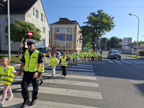 Na zdjęciu widoczne dzieci, które przez jednię po przejściu dla pieszych przeprowadzają policjanci.
